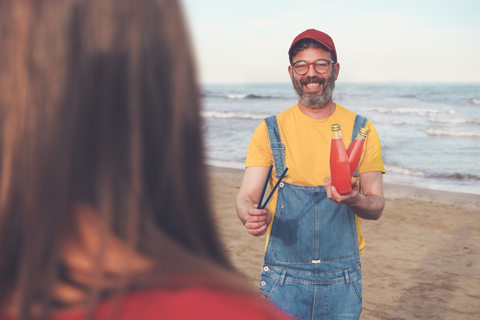 Glücklicher Mann in Latzhose am Strand, der einer Frau ein Erfrischungsgetränk anbietet, lizenzfreies Stockfoto
