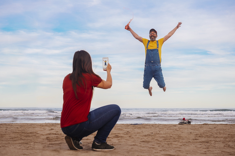 Frau macht Smartphone-Foto von glücklichem Mann, der am Strand springt, lizenzfreies Stockfoto