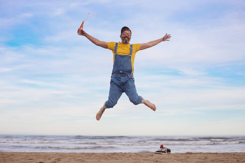 Porträt eines glücklichen Mannes in Latzhose, der mit einer Limonade in der Hand am Strand herumspringt - RTBF01186