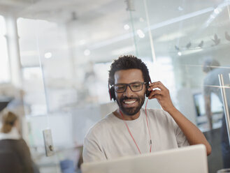 Lächelnder kreativer Geschäftsmann mit Kopfhörern bei der Arbeit am Laptop im Büro - HOXF03430