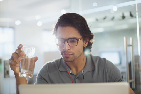 Konzentrierter Geschäftsmann mit Kopfhörern, der am Laptop Wasser trinkt, lizenzfreies Stockfoto