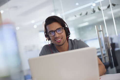 Portrait lächelnder, selbstbewusster kreativer Geschäftsmann mit Kopfhörern am Laptop im Büro - HOXF03459