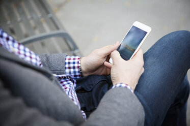 Mittelteil eines Mannes, der ein Mobiltelefon benutzt, während er auf einer Bank im Park sitzt - CAVF45714