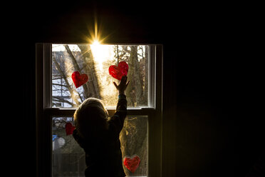 Rear view of boy sticking heart shapes on window at home - CAVF46041