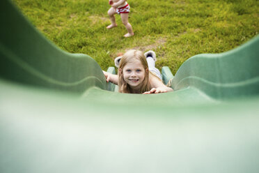 Porträt eines glücklichen Mädchens, das rutscht, während sein Bruder auf dem Spielplatz spielt - CAVF46066