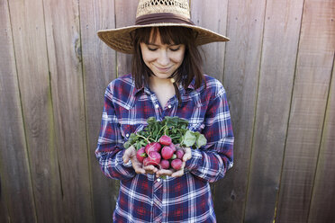 Smiling young female farmer holding radishes while standing against wooden fence - CAVF46203