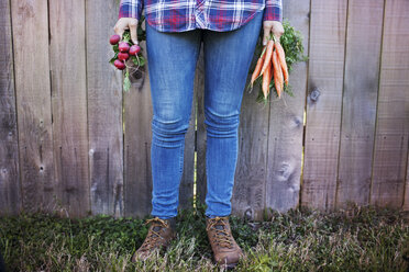Low section of female farmer holding vegetables while standing against fence at organic farm - CAVF46205