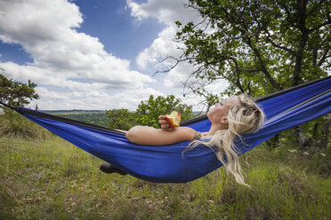 Frau hält einen Apfel in der Hand, während sie im Urlaub auf einem Feld in der Hängematte liegt - CAVF46233