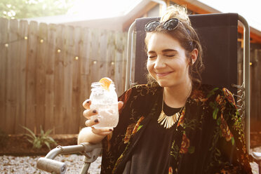 Happy woman with eyes closed holding drink jar while sitting in yard - CAVF46289