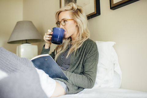 Frau mit Tagebuch, die zu Hause auf dem Bett sitzt und etwas trinkt, lizenzfreies Stockfoto
