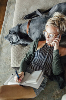 Draufsicht auf eine Frau, die sich mit einem Smartphone über ihr Tagebuch unterhält, während sie sich auf ihrer Dogge zu Hause entspannt - CAVF46348