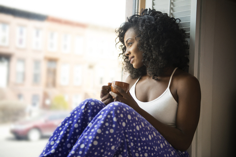 Lächelnde Frau, die durch das Fenster schaut, während sie zu Hause Kaffee trinkt, lizenzfreies Stockfoto