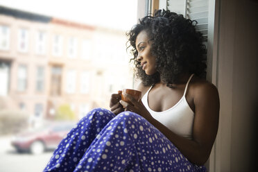 Lächelnde Frau, die durch das Fenster schaut, während sie zu Hause Kaffee trinkt - CAVF46392