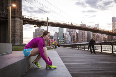 Woman tying shoe lace while sitting on steps against Brooklyn bridge - CAVF46459