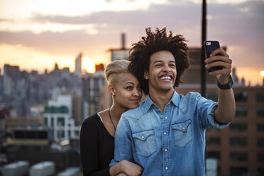 Glückliches Paar, das ein Selfie macht, während es bei Sonnenuntergang auf dem Dach steht - CAVF46495