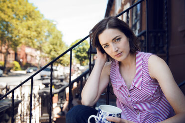Porträt einer Frau, die eine Kaffeetasse hält, während sie auf einer Treppe vor einem Gebäude sitzt - CAVF46554