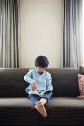 Boy reading book while sitting on sofa against wall at home - CAVF46562