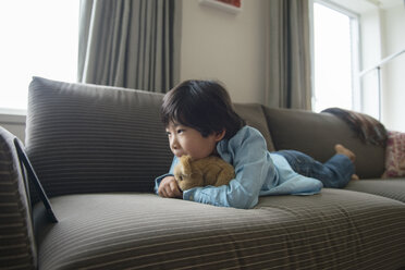 Boy looking at tablet while lying with stuffed toy on sofa at home - CAVF46570