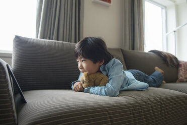 Junge schaut auf ein Tablet, während er mit einem Stofftier zu Hause auf dem Sofa liegt - CAVF46570