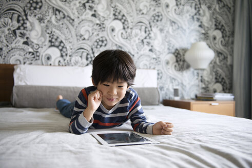 Glücklicher Junge, der ein digitales Tablet benutzt, während er auf dem Bett liegt - CAVF46582