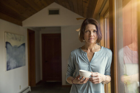 Porträt einer Frau, die eine Kaffeetasse in der Hand hält, während sie am Fenster im Wohnzimmer lehnt - CAVF46607