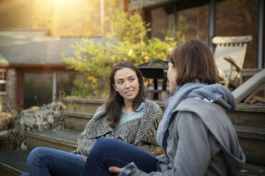 Smiling daughter and mother talking while sitting on steps outside log cabin in forest - CAVF46626