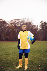 Happy confident soccer player holding ball while standing on playing field against clear sky - CAVF46647