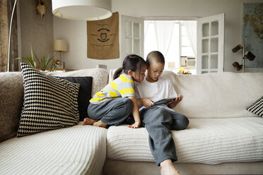 Sister looking at brother using tablet computer while sitting on sofa in living room - CAVF46704