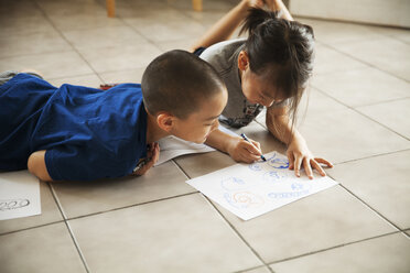 Brother looking at sister drawing on paper while lying on floor in living room - CAVF46712