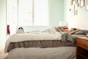 Katze betrachtet schlafende Frau auf dem Bett vor dem Fenster zu Hause - CAVF46742