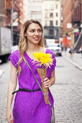 Lächelnde Frau, die einen Blumenstrauß hält, während sie auf einer gepflasterten Straße in der Stadt spazieren geht - CAVF46786