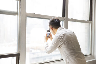 Businessman photographing from window through smart phone in creative office - CAVF46813