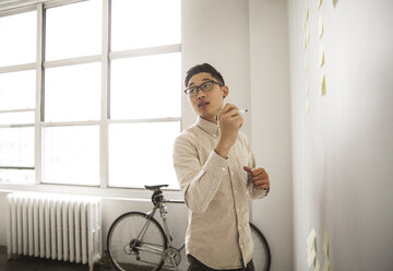 Confident businessman making sticky notes standing against wall in creative office - CAVF46815