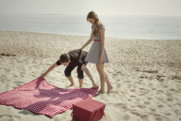 Freundin betrachtet Mann, der eine Picknickdecke auf den Sand am Strand legt - CAVF46826
