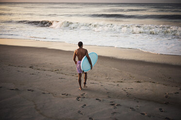 Rückansicht eines Mannes, der ein Surfbrett trägt, während er auf dem Sand am Strand läuft - CAVF46840