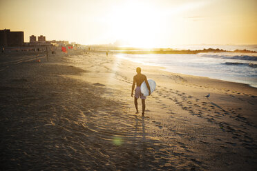 Mann in voller Länge, der ein Surfbrett trägt, während er auf dem Sand am Strand läuft - CAVF46842