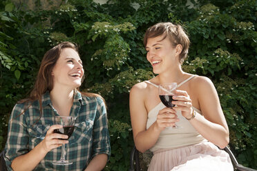 Happy friends enjoying wine while sitting on chair in yard - CAVF46856