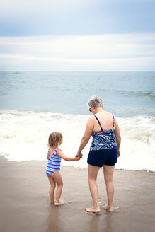 Granddaughter and grandmother holding hands while standing on shore at beach - CAVF46876