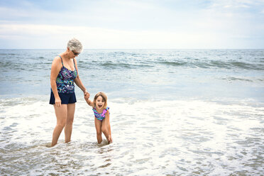 Verspieltes Mädchen mit Großmutter im Wasser stehend am Strand - CAVF46878