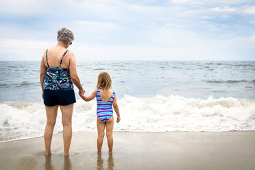 Rear view of granddaughter and grandmother standing on shore at beach - CAVF46880