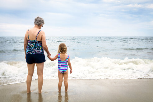 Rear view of granddaughter and grandmother standing on shore at beach - CAVF46880