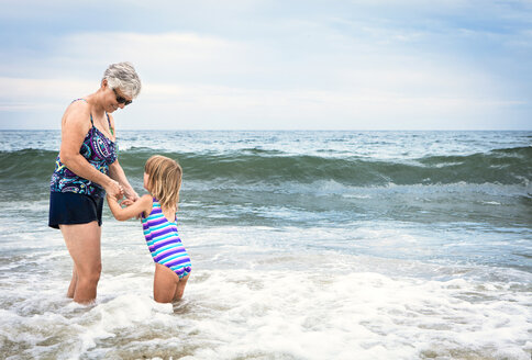 Playful grandmother with granddaughter standing in water at beach - CAVF46881