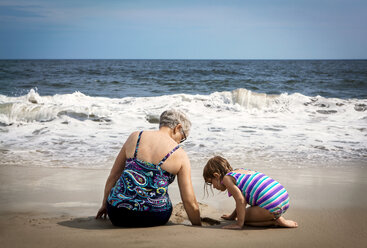 Girl looking down while grandmother digging in sand at beach - CAVF46885