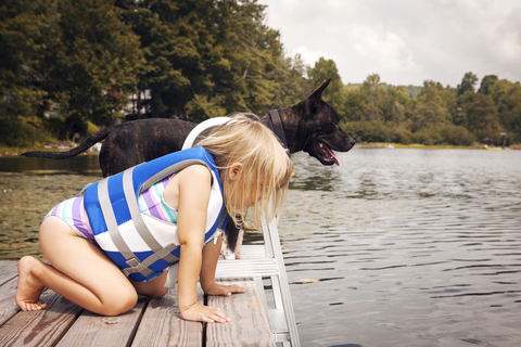 Seitenansicht eines Mädchens, das nach unten schaut, während es neben einem Hund auf einem Pier kniet, lizenzfreies Stockfoto