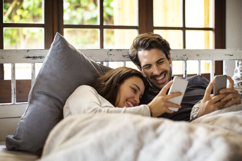 Ein glückliches Paar benutzt sein Smartphone, während es zu Hause auf dem Bett liegt - CAVF47048