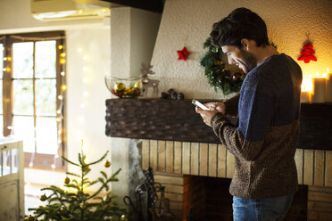 Smiling man using smart phone while standing at home during Christmas - CAVF47069