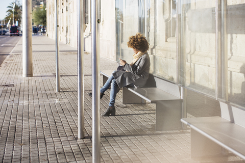 Frau benutzt Smartphone, während sie auf dem Sitz an der Bushaltestelle sitzt, lizenzfreies Stockfoto
