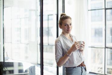 Porträt einer Geschäftsfrau, die ein Getränk zu sich nimmt und sich im Büro ans Fenster lehnt - CAVF47212