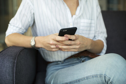 Mittelteil einer Geschäftsfrau, die ein Mobiltelefon benutzt, während sie auf dem Sofa im Büro sitzt - CAVF47213