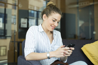 Lächelnde Geschäftsfrau, die ein Mobiltelefon benutzt, während sie auf dem Sofa im Büro sitzt - CAVF47214